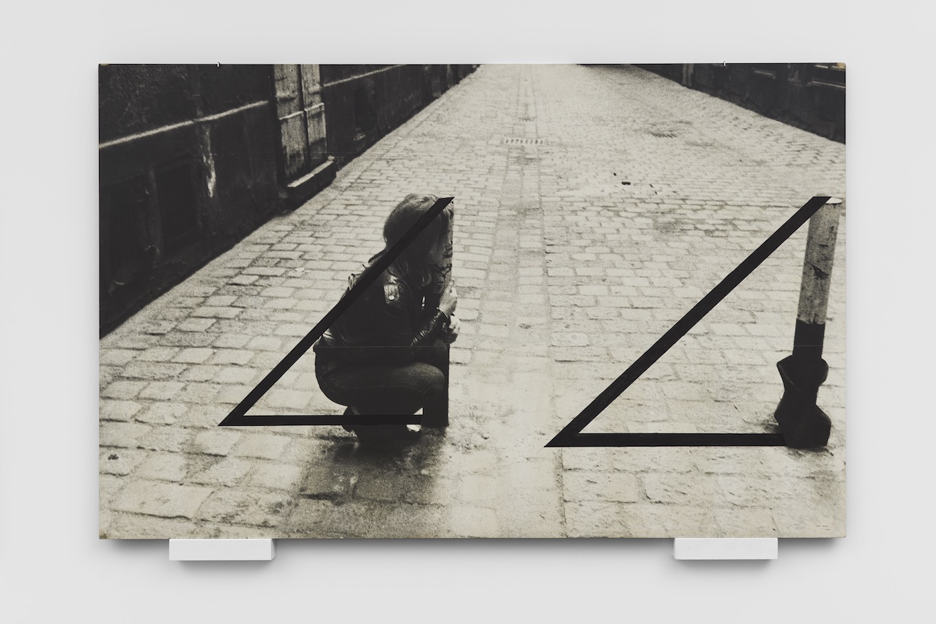 Black and white image of a woman crouching next to two traffic poles, holding onto one to form the shape of a triangle, which is outlined in black next to both traffic poles