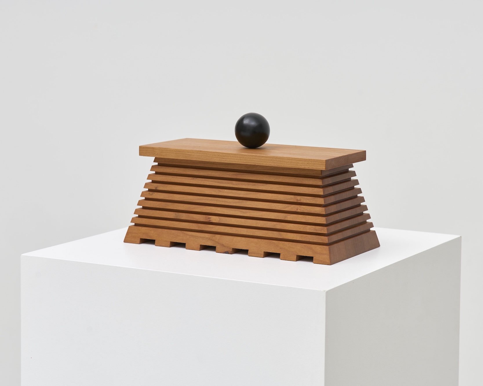 Ryan Preciado’s “Atentamente,” a small grooved rectangular box made of wood with spherical black lid on a white plinth