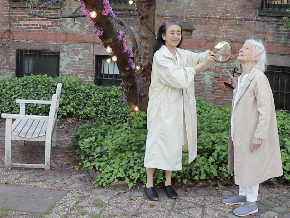 Otake and Jonas standing together on a patio as Otake holds a mirror up to Jonas's face