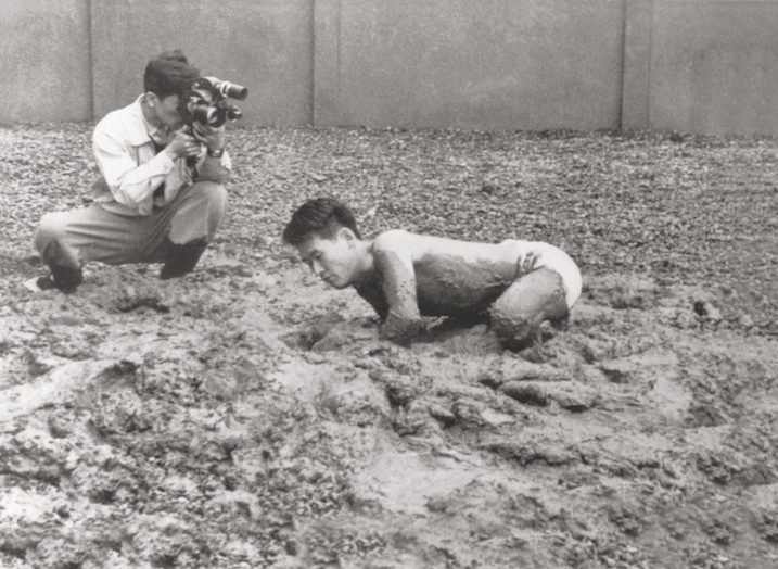 A black and white photo of a man crawling in mud with someone photographing him