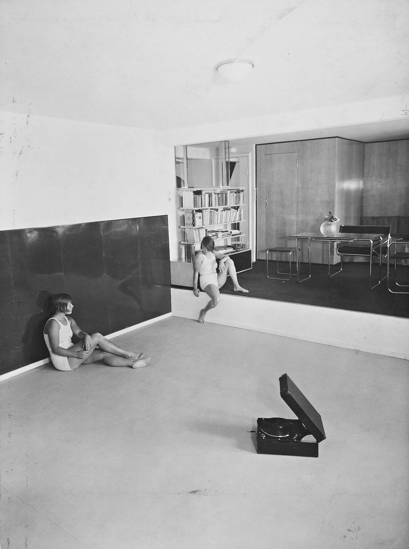 Black and white wide shot of two women in athletic clothing sitting in an apartment with a gym in it