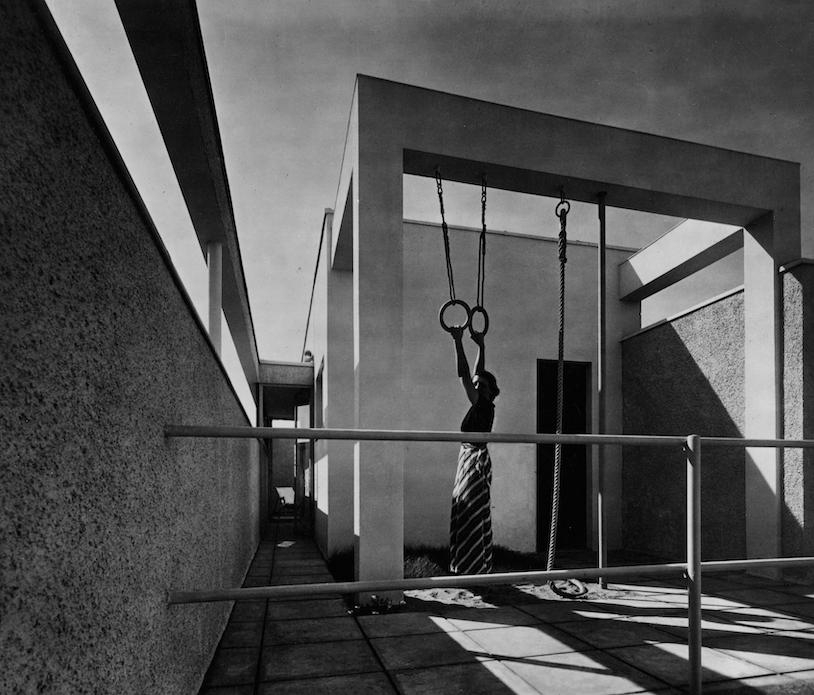 Black and white wide shot of Gege Figini holding onto gymnastics rings that are hanging from a small gymnasium on her house's rooftop terrace