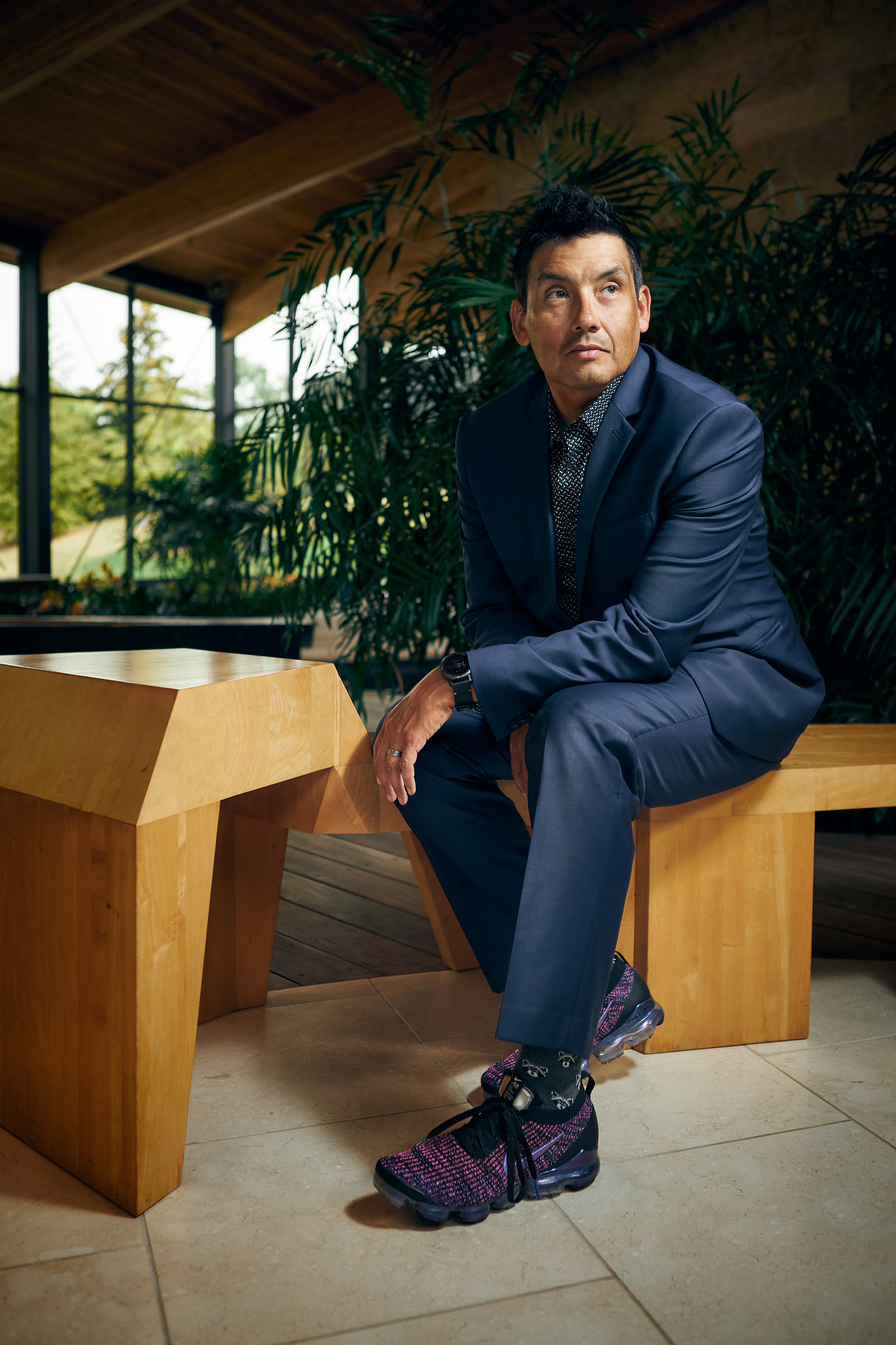Portrait of Chris Cornelius in suit and sneakers sitting on wooden bench inside a house