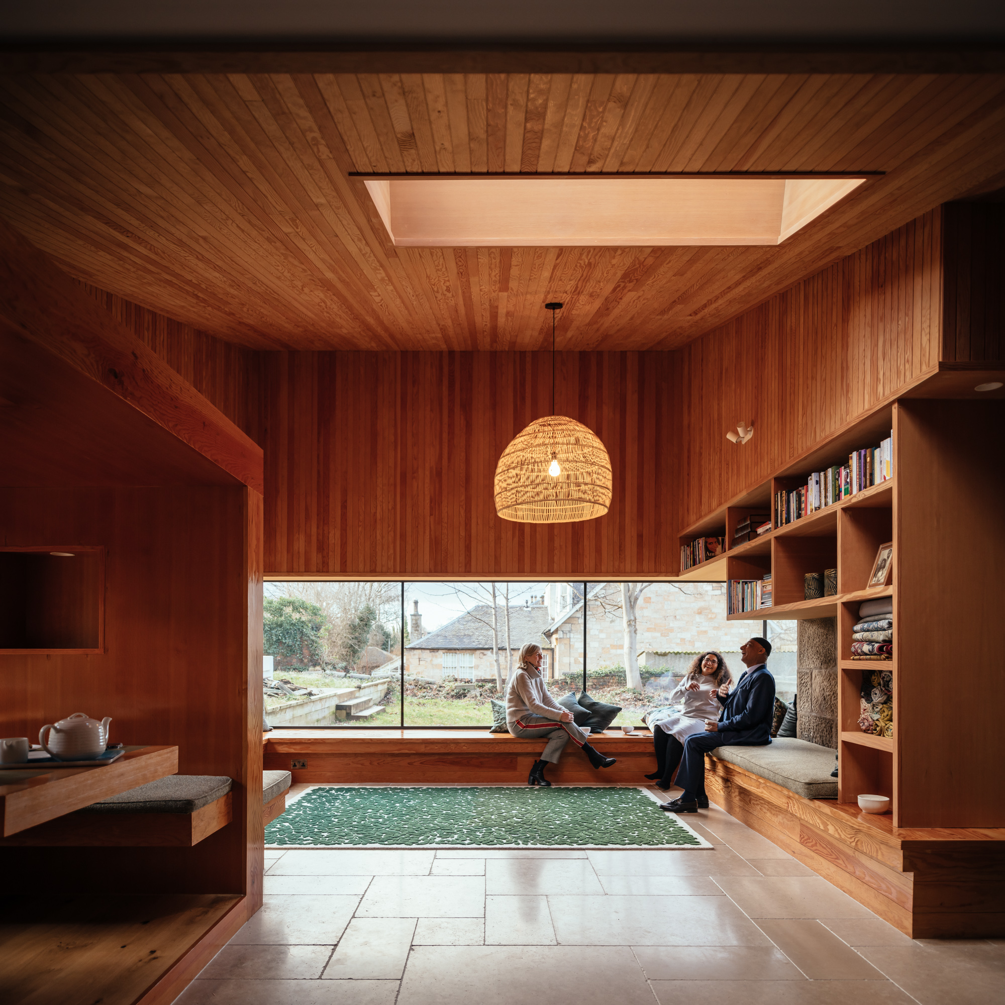 Three adults sitting and talking in a large corner window seat of a room with wooden walls and ceiling