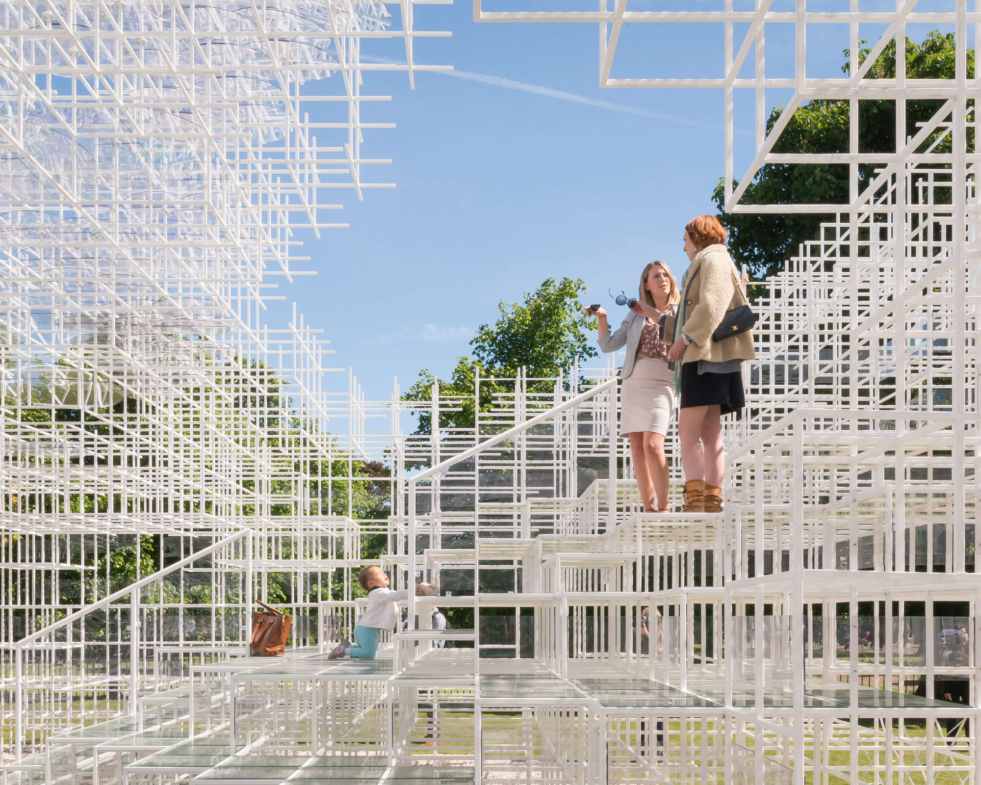 Large 3-D grid structure of thin white bars with two women standing on it talking and a baby below them climbing on it